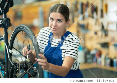 Professional young girl master wearing uniform fixing bike, lubricating bicycle chain in sport store Professional young girl master wearing uniform fixing bike, lubricating bicycle chain in sport store 114146505