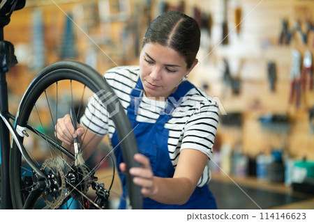 Professional young girl master wearing uniform fixing bike, lubricating bicycle chain in sport store 114146623