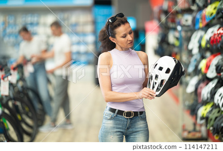 Attentive sporty woman buyer choosing safe bicycle helmet in sports store Attentive sporty woman buyer choosing safe bicycle helmet in sports store 114147211