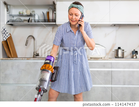 Woman in dress and headband is vacuuming floor and talking on mobile phone, ordering food delivery Woman in dress and headband is vacuuming floor and talking on mobile phone, ordering food delivery 114147437