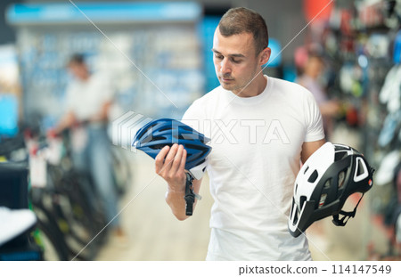 Focused cyclist choosing new bicycle helmet in sports store 114147549