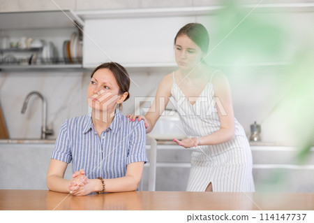 Offended mother sitting at the kitchen table, daughter standing behind 114147737