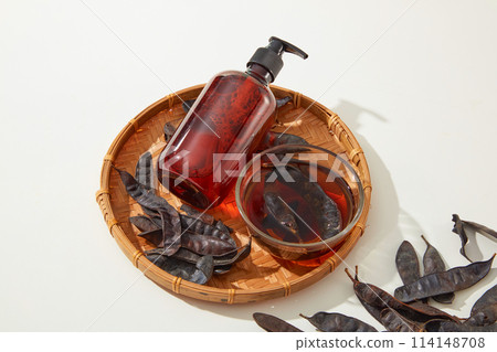 A rattan tray contains a blank label bottle, a glass bowl of brown liquid and some black locust pods isolated on white background. High angle view with blank space for adding elements 114148708