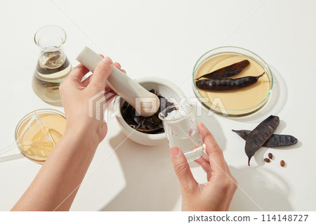 A hand is holding a glass of water ready for pouring into a mortar, process of producing manufacturing made from black locust pod. Top view photo with blank space for design 114148727