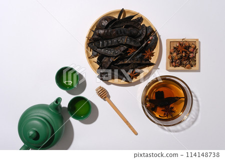 Organic beverage concepts with a green tea pot and two tea cups placed by a wooden dish of black locust, a wooden tray with herbs above and a glass bowl with brown liquid. Space for text, top view Organic beverage concepts with a green tea pot and two tea cups placed by a wooden dish of black locust, a wooden tray with herbs above and a glass bowl with brown liquid. Space for text, top view 114148738