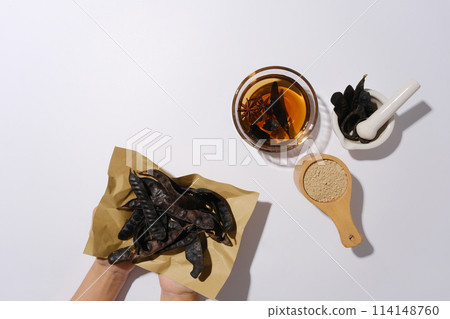 Beautiful hand holding a vintage paper with locust fruits above, a bowl of locust liquid, a cup of brown powder, mortar and pestle, white background. Space for displaying Beautiful hand holding a vintage paper with locust fruits above, a bowl of locust liquid, a cup of brown powder, mortar and pestle, white background. Space for displaying 114148760