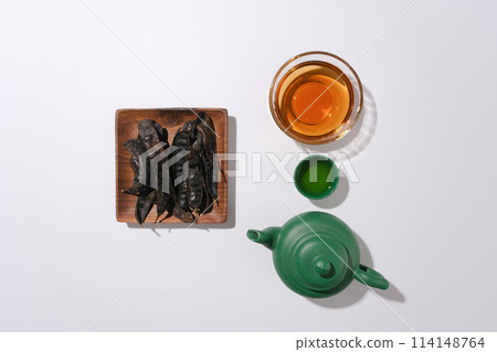 Layout for drinks with a rectangle wooden dish contains black locust pods, a green tea pot, tea cup and a glass bowl within brown fluid, flat lay on white table. Blank space, top view photo Layout for drinks with a rectangle wooden dish contains black locust pods, a green tea pot, tea cup and a glass bowl within brown fluid, flat lay on white table. Blank space, top view photo 114148764