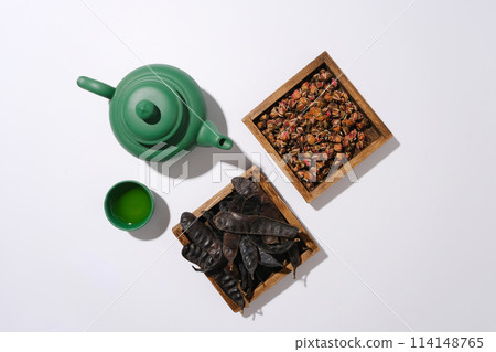 Top view photo with a green tea pot, a cup of tea, wooden tray of dried rose buds and a tray of black locust fruits placed side by side on white background. Space for text or designing Top view photo with a green tea pot, a cup of tea, wooden tray of dried rose buds and a tray of black locust fruits placed side by side on white background. Space for text or designing 114148765