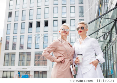 two businesswomen standing outside with coffee with buildings on background two businesswomen standing outside with coffee with buildings on background 114149084