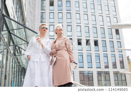 two businesswomen standing outside with coffee with buildings on background two businesswomen standing outside with coffee with buildings on background 114149085