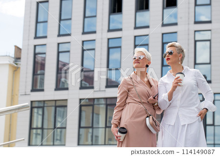 two businesswomen standing outside with coffee with buildings on background two businesswomen standing outside with coffee with buildings on background 114149087