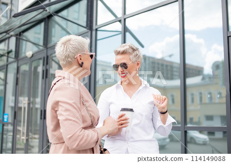 two businesswomen standing outside with coffee with buildings on background two businesswomen standing outside with coffee with buildings on background 114149088