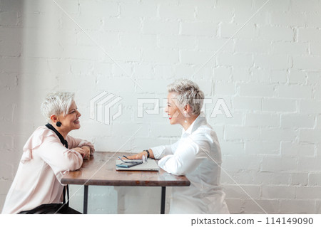 Two female senior collegues sitting next to each other in an cafe Two female senior collegues sitting next to each other in an cafe 114149090