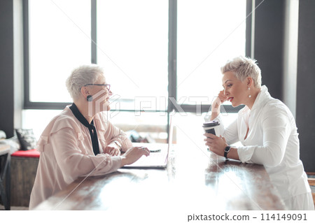 Two female senior collegues sitting next to each other in an cafe Two female senior collegues sitting next to each other in an cafe 114149091