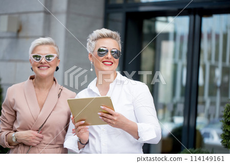 Two businesswoman walking on street near building. business woman going together with tablet computer Two businesswoman walking on street near building. business woman going together with tablet computer 114149161