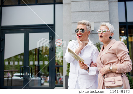 Two businesswoman walking on street near building. business woman going together with tablet computer Two businesswoman walking on street near building. business woman going together with tablet computer 114149173