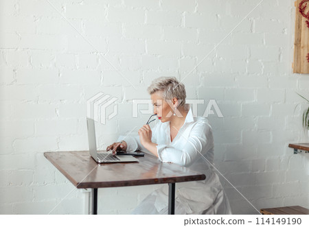 Smiling businesswoman is sitting at table with laptop and using think about business Smiling businesswoman is sitting at table with laptop and using think about business 114149190