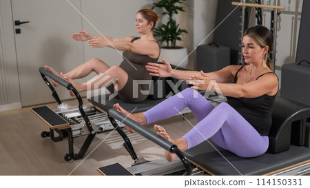 Two pregnant women do Pilates exercises on a reformer. Yoga class for pregnant women. Two pregnant women do Pilates exercises on a reformer. Yoga class for pregnant women. 114150331