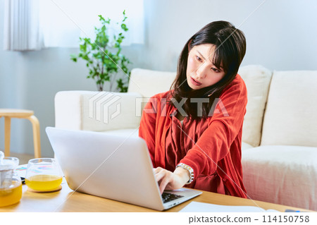 Busy woman making calls on her smartphone while working remotely on a laptop in the living room at home 114150758