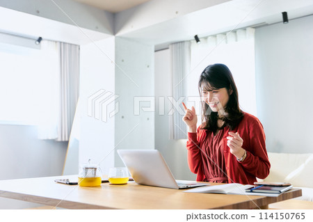 A smiling Japanese woman in her 30s gesturing to a video call partner at her desk in the living room of her home 114150765