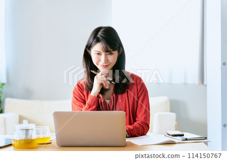 客廳桌上，一名30多歲的女士拿著筆，看著筆記型電腦，正在思考。 114150767