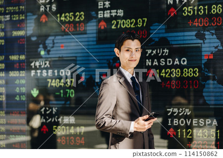 一名 30 多歲的男子站在顯示世界股票價格的電子公告牌前，微笑著看著鏡頭。 114150862