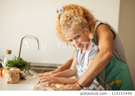 Beautiful young mom and her cute little daughter rolling out dough at home. 114153449