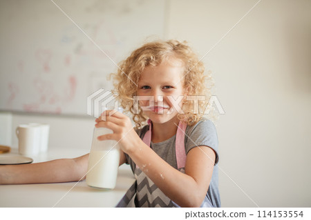 Cute little girl with fair frizzy hair drinking a glass of milk in kitchen 114153554
