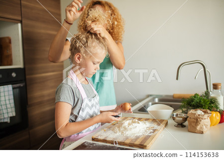 Little girl learn to make dough and her caring mother tying her hair up. Little girl learn to make dough and her caring mother tying her hair up. 114153638
