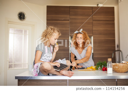 Little girl and her daughter baking cookies at home, reading recipe on laptop. Little girl and her daughter baking cookies at home, reading recipe on laptop. 114154256