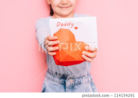Little girl holding a drawn card and a heart for Father's Day in front of a pink background 114154291