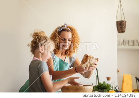 Little girl and her mother sprinkling flour into a bowl and smiling while baking Little girl and her mother sprinkling flour into a bowl and smiling while baking 114154382