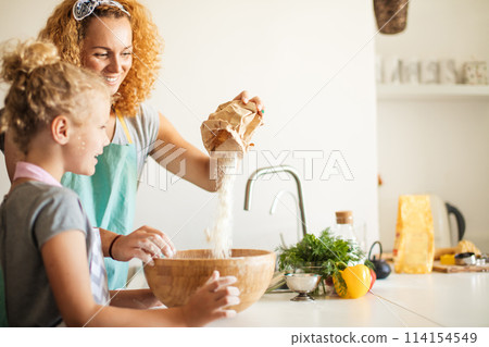 Little girl and her mother sprinkling flour into a bowl and smiling while baking Little girl and her mother sprinkling flour into a bowl and smiling while baking 114154549
