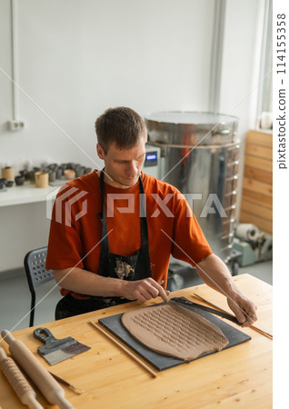 A potter cuts a piece of rolled clay with patterns. Vertical photo. A potter cuts a piece of rolled clay with patterns. Vertical photo. 114155358