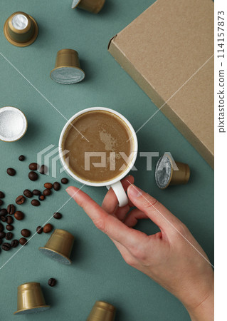 Coffee capsules and beans, cup with coffee drink in hand and box on green background, top view Coffee capsules and beans, cup with coffee drink in hand and box on green background, top view 114157873