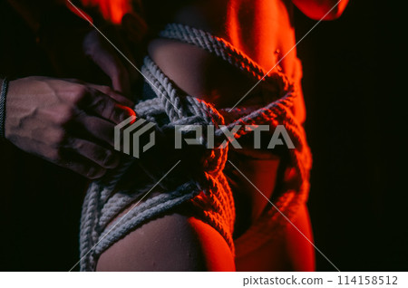 Man tightens ropes using Japanese shibari technique on woman's body in red light.  114158512