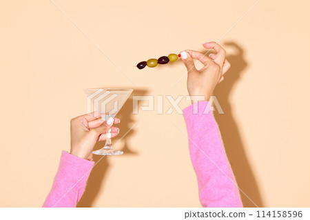 Female hand holding martini glass with a green olive on a stick against light background. Female hand holding martini glass with a green olive on a stick against light background. 114158596
