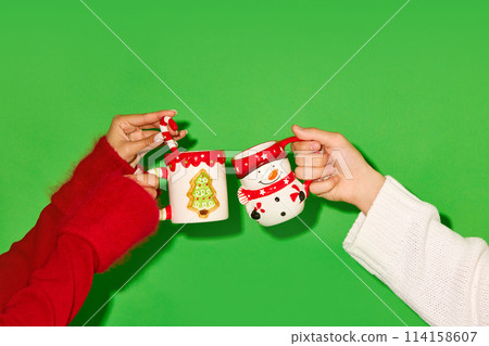 Hands holding mugs decorated in Christmas theme against green background. Hot tea, cocoa and hot chocolate 114158607