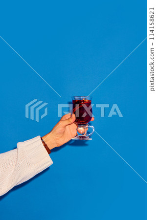 Hand holding glass with mulled wine made with red wine and cinnamon against blue background. Traditional winter drink Hand holding glass with mulled wine made with red wine and cinnamon against blue background. Traditional winter drink 114158621