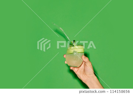 Hand holding glass with refreshing sweet and sour cocktail with lien and cucumber taste against green background Hand holding glass with refreshing sweet and sour cocktail with lien and cucumber taste against green background 114158656
