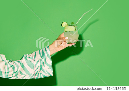 Female hand holding glass with refreshing sweet and sour cocktail with lien and cucumber taste against green background Female hand holding glass with refreshing sweet and sour cocktail with lien and cucumber taste against green background 114158665