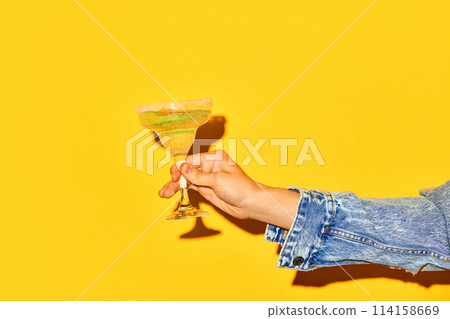Male hand holding sweet and sour margarita cocktail with slice of orange against bright yellow background Male hand holding sweet and sour margarita cocktail with slice of orange against bright yellow background 114158669