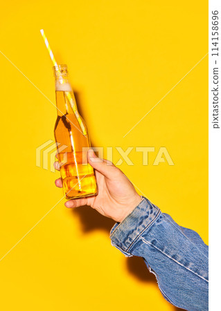 Human hand holding bottle with lemonade with straw against bright yellow background Human hand holding bottle with lemonade with straw against bright yellow background 114158696