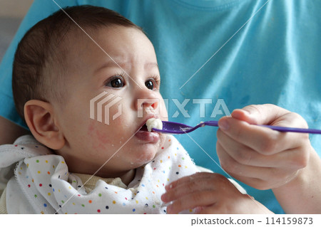 Portrait of little baby boy eating food. Baby with a spoon in feeding chair. Cute baby eating first meal Portrait of little baby boy eating food. Baby with a spoon in feeding chair. Cute baby eating first meal 114159873