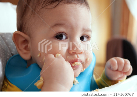 Portrait of little baby boy eating food. Baby with a spoon in feeding chair. Cute baby eating first meal 114160767
