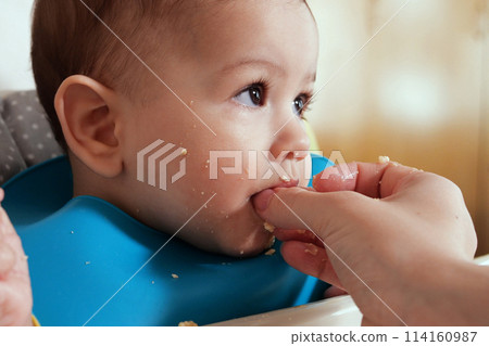 Portrait of little baby boy eating food. Baby with a spoon in feeding chair. Cute baby eating first meal 114160987