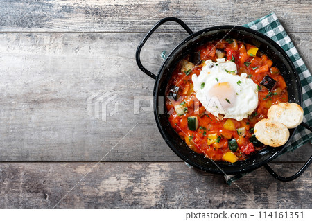 Vegetable pisto manchego with tomatoes, zucchini, peppers, onions,eggplant and egg, served in frying pan on wooden table 114161351