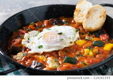 Vegetable pisto manchego with tomatoes, zucchini, peppers, onions,eggplant and egg, served in frying pan on wooden table 114161355