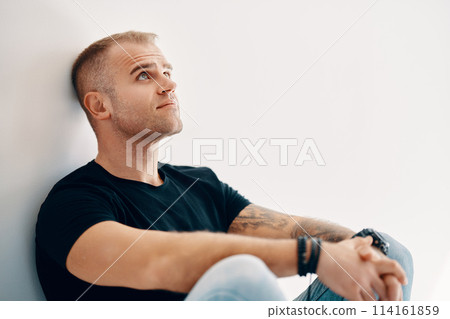 Portrait of handsome thoughtful man sitting on floor leaning to the wall Portrait of handsome thoughtful man sitting on floor leaning to the wall 114161859