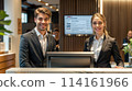 Two people are standing at a reception desk, smiling for the camera. The woman is wearing a black jacket and the man is wearing a suit 114161966